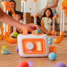 A child's hand pushes a colorful toy cube on a wooden floor, with children and adults sitting in the background playing with a large orange and clear toy maze.