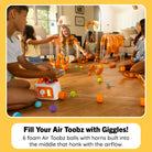 A group of children playing with an orange and clear foam air toy set on a hardwood floor in a living room. The toy includes curved clear tubes, orange connectors, and several colorful air-toz balls, with one child using a small white and orange air pump.
