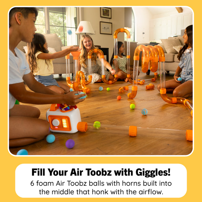 A group of children playing with an orange and clear foam air toy set on a hardwood floor in a living room. The toy includes curved clear tubes, orange connectors, and several colorful air-toz balls, with one child using a small white and orange air pump.