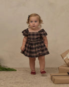 A toddler girl stands in a studio wearing a dark plaid dress with puff sleeves and burgundy shoes, posed against a textured beige backdrop with wrapped gifts nearby.