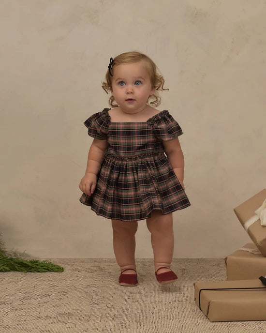A toddler girl stands in a studio wearing a dark plaid dress with puff sleeves and burgundy shoes, posed against a textured beige backdrop with wrapped gifts nearby.