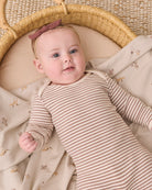 A baby lying in a woven bassinet, wearing a beige and brown striped long-sleeve bodysuit with a dusty rose bow headband.