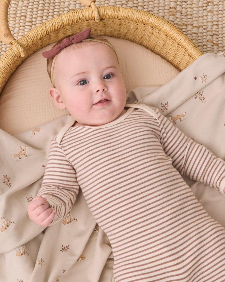 A baby lying in a woven bassinet, wearing a beige and brown striped long-sleeve bodysuit with a dusty rose bow headband.