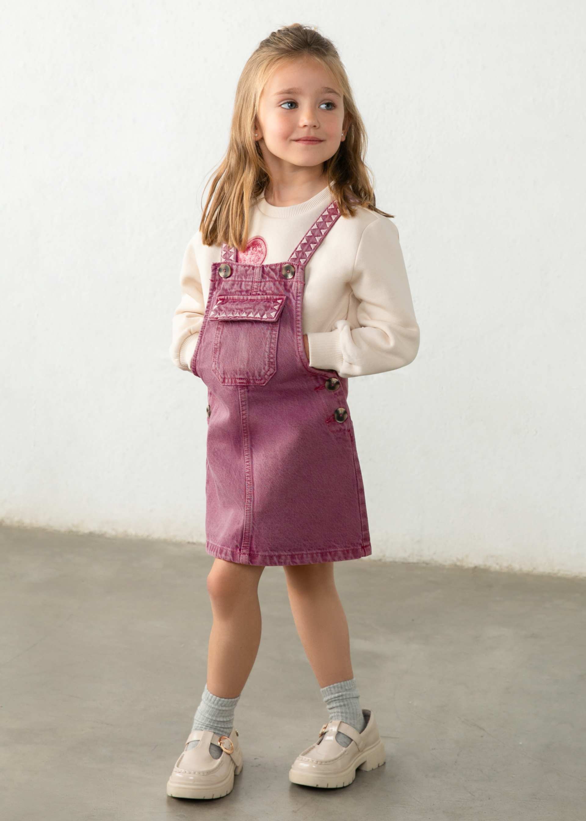 A child wearing dusty rose denim overall with pink embroidered details and a cream sweatshirt underneath, shown from the chest up against a light neutral background.