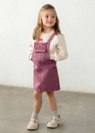 A child wearing dusty rose denim overall with pink embroidered details and a cream sweatshirt underneath, shown from the chest up against a light neutral background.