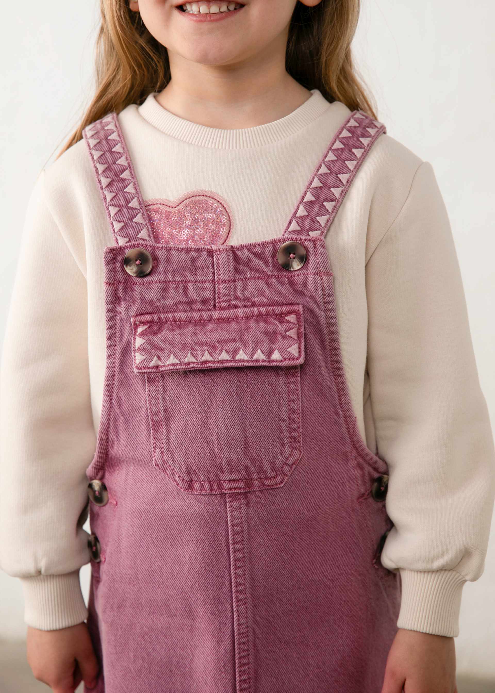 A child wearing dusty rose denim overall with pink embroidered details and a cream sweatshirt underneath, shown from the chest up against a light neutral background.