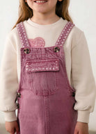 A child wearing dusty rose denim overall with pink embroidered details and a cream sweatshirt underneath, shown from the chest up against a light neutral background.