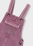 A dusty pink denim overall dress for kids, featuring adjustable shoulder straps with a zigzag pattern, side pockets with metal snap buttons, and a front bib pocket, shown against a plain white background.