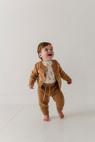 A baby wearing a matching brown knit cardigan and pants set over a white patterned top, standing barefoot on a white studio floor.