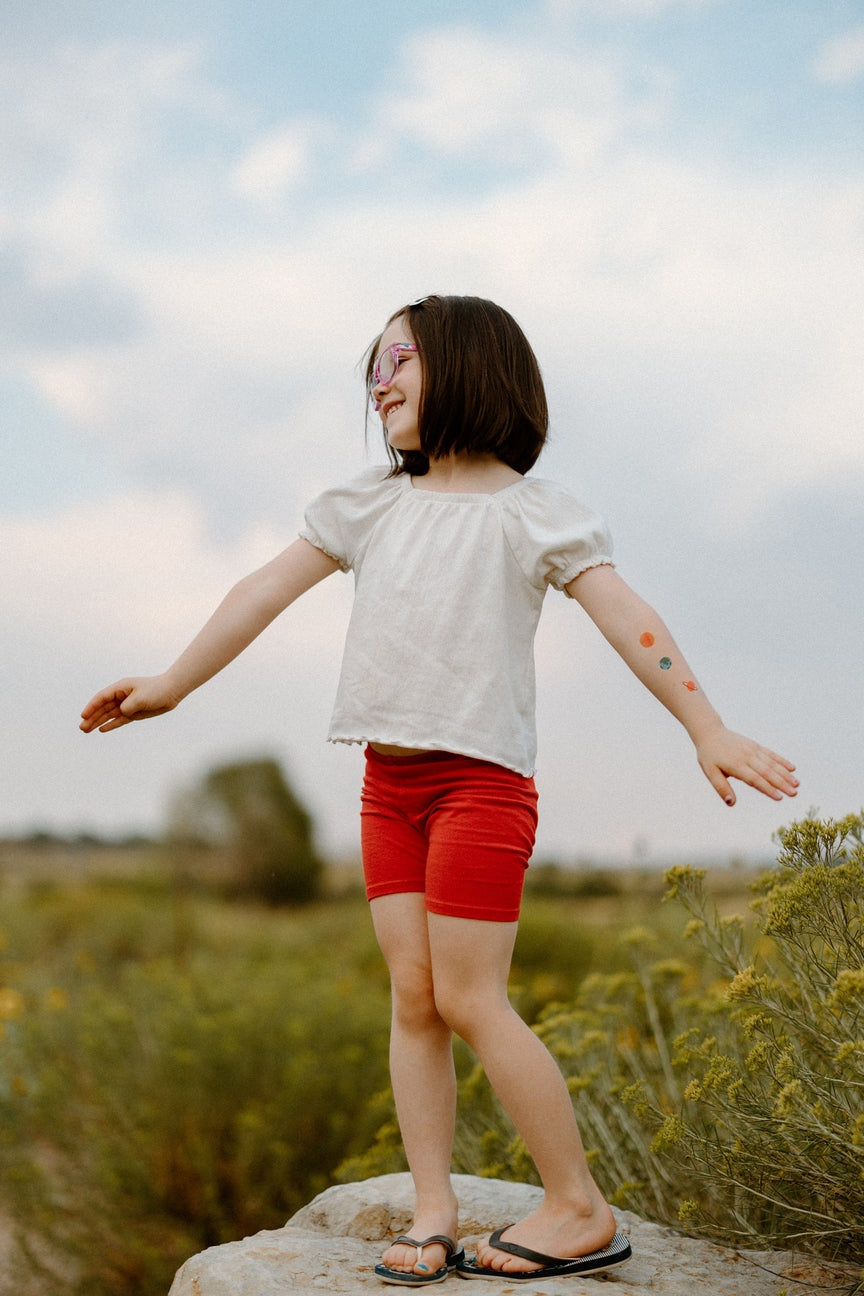 Child standing on a rock in a field with arms outstretched, wearing a white shirt and red shorts.