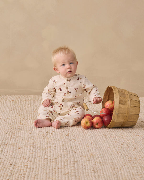 A baby sitting on a textured beige rug next to a wooden basket tipped over with red apples, wearing a cream-colored long-sleeve romper with a small brown floral pattern.
