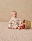 A baby sitting on a textured beige rug next to a wooden basket tipped over with red apples, wearing a cream-colored long-sleeve romper with a small brown floral pattern.
