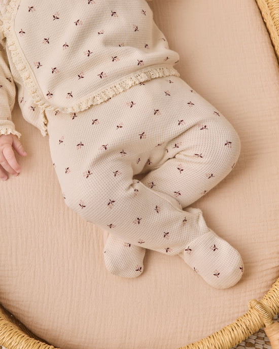A baby wearing a cream-colored waffle-knit footed sleeper with a small brown floral pattern and lace trim, lying on a beige muslin blanket inside a woven basket.