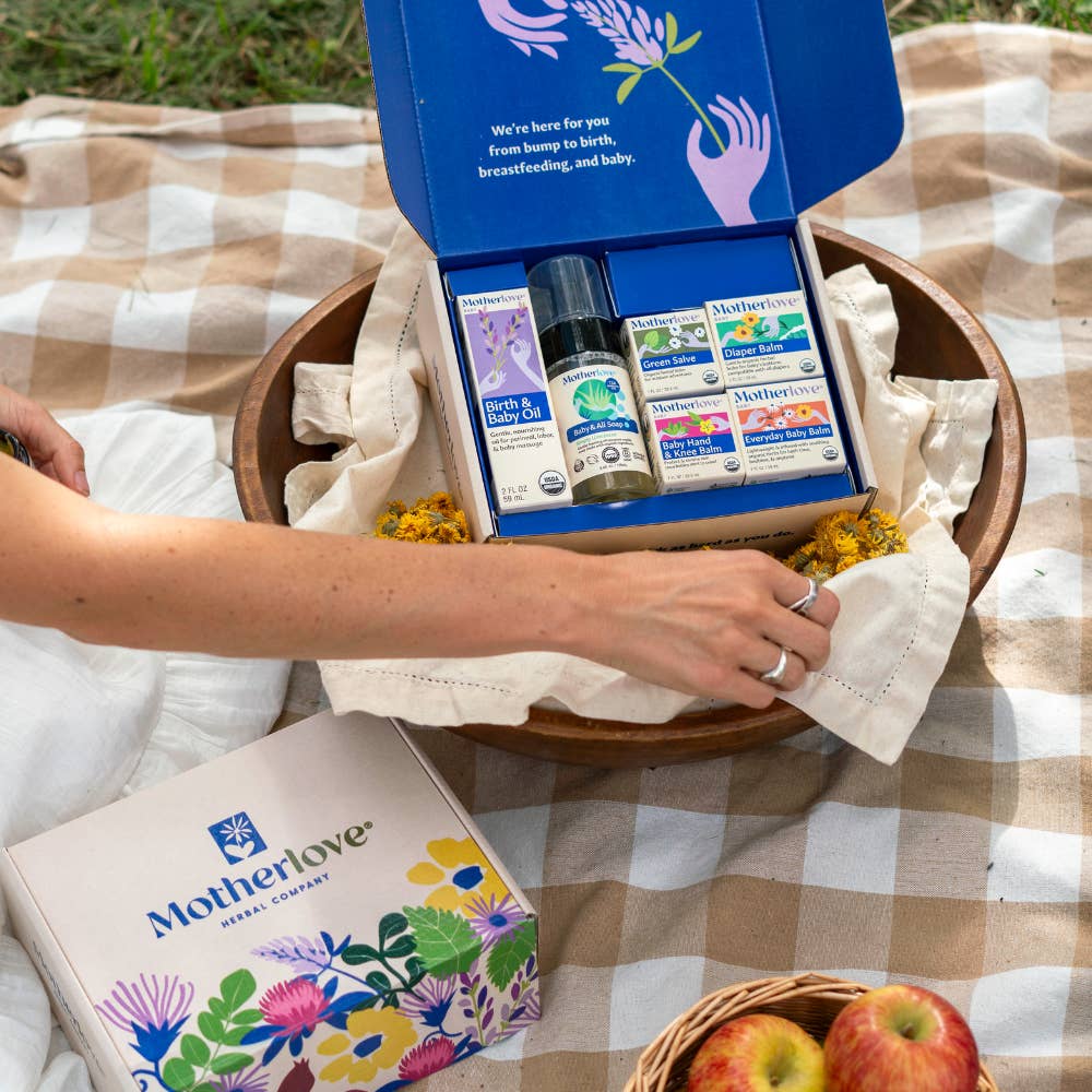 Open box of Motherlove products on a checkered blanket with apples and flowers.