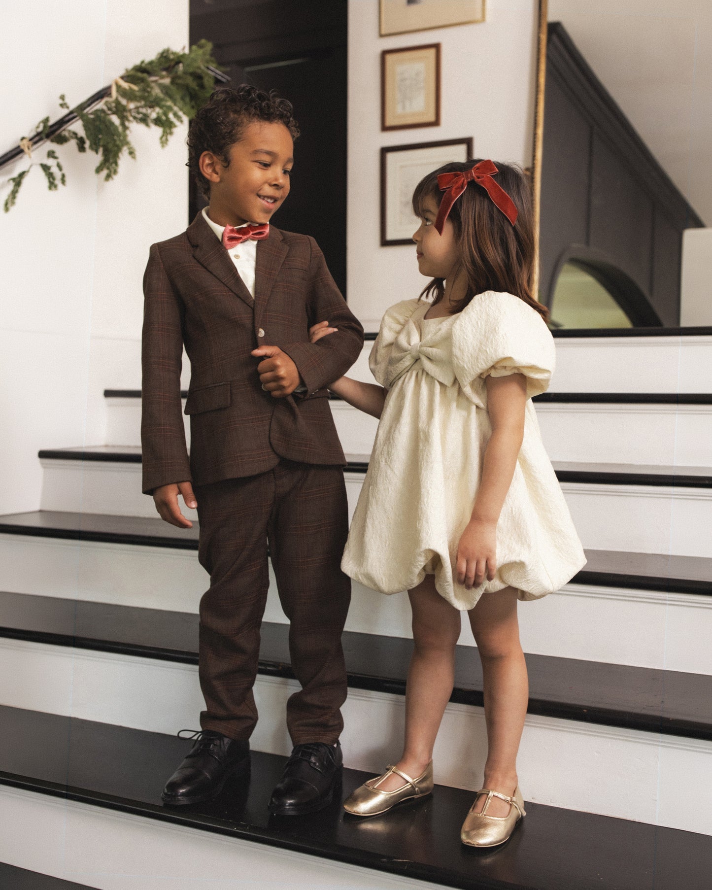 A young boy in a brown textured suit with a red bow tie stands next to a girl in a cream puff-sleeve dress with a red bow headband, posing on a black and white staircase.