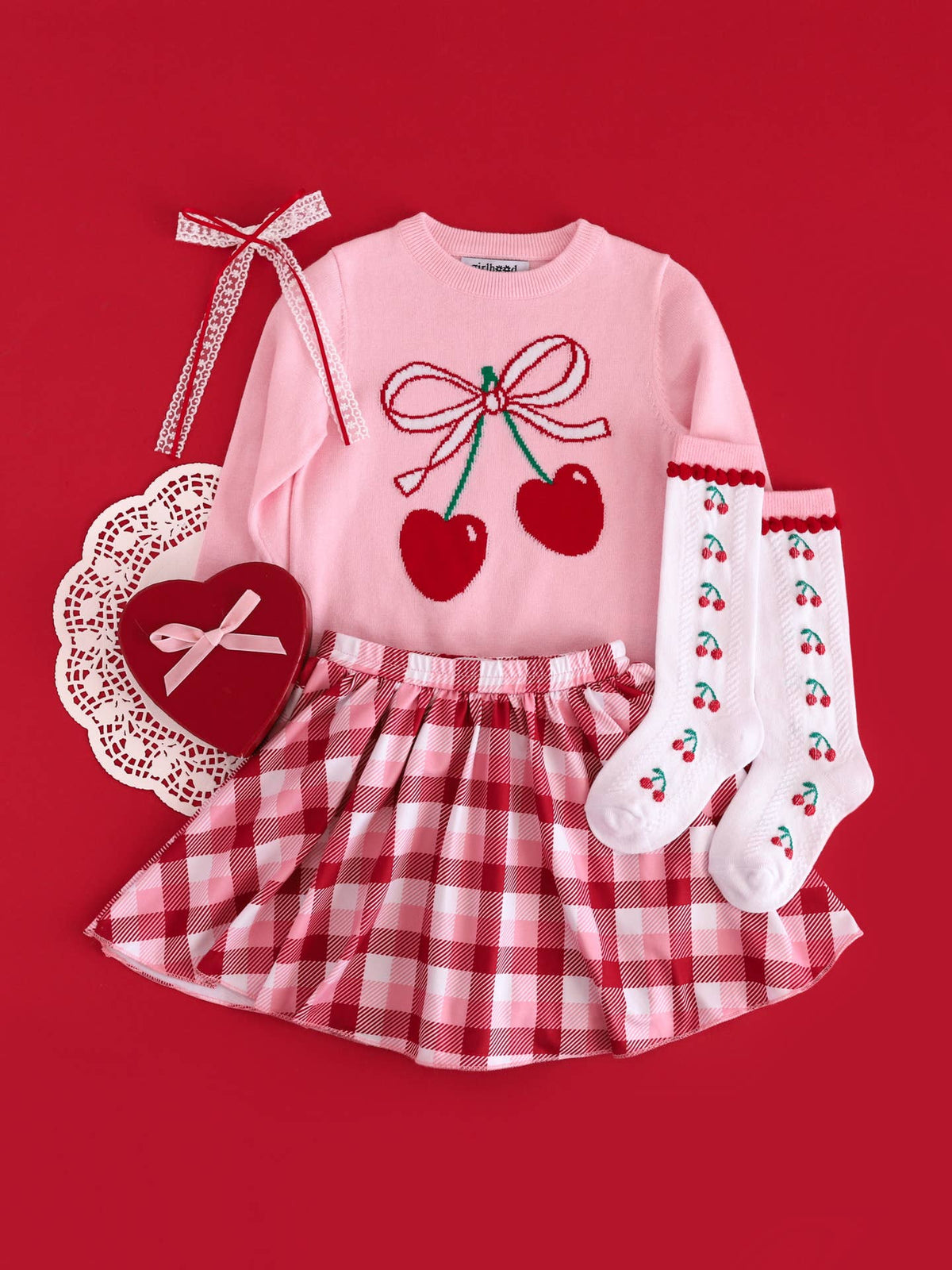 Children's outfit with pink top, red plaid skirt, white socks with red cherries, and decorative heart on a red background.