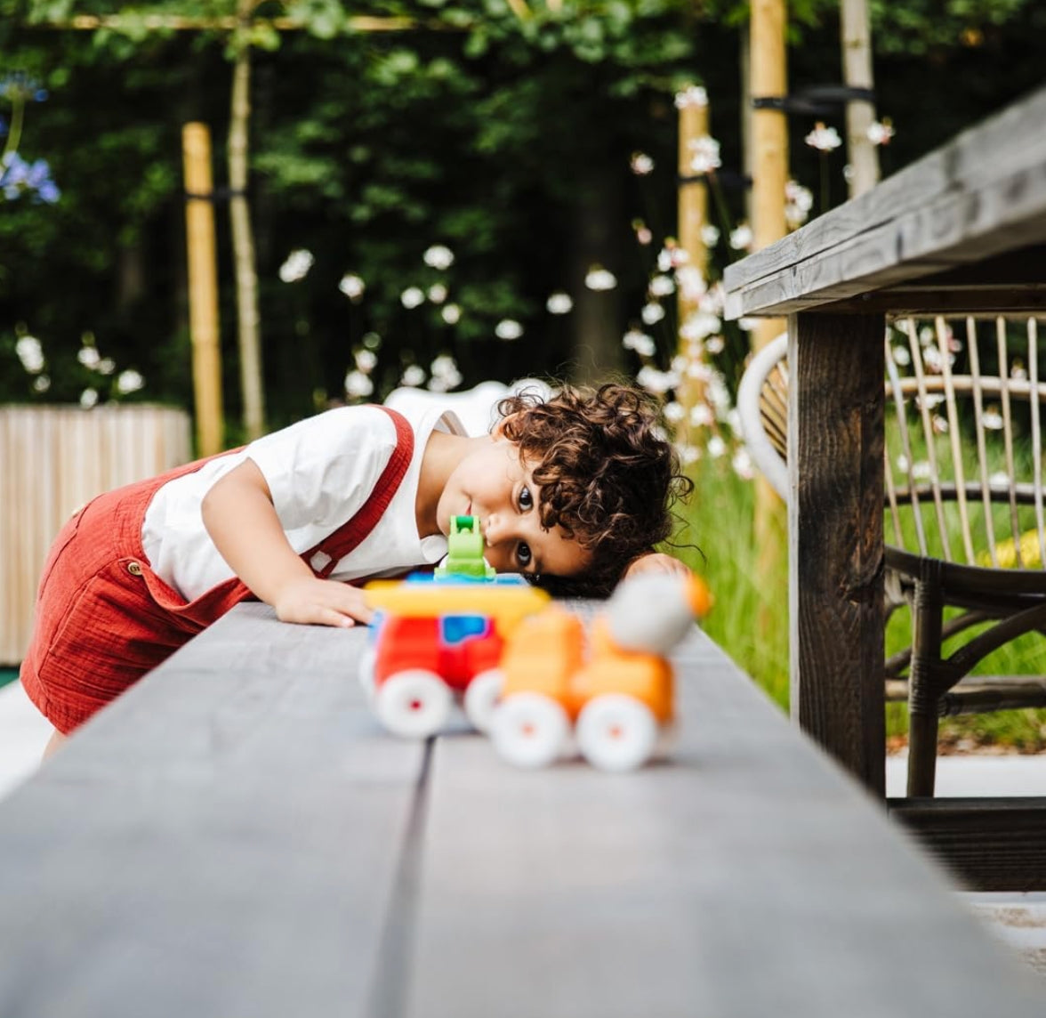 A young child with curly brown hair leans over a wooden table in a garden setting, looking at a small toy train. The child is wearing a white t-shirt and red overalls.