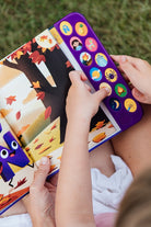 A child holding open "What Does Ñ Sound Like?: A Spanish-English Sound Book," showing an interior illustrated page with a purple animated character resembling the letter Ñ, set against an autumn landscape with falling leaves.