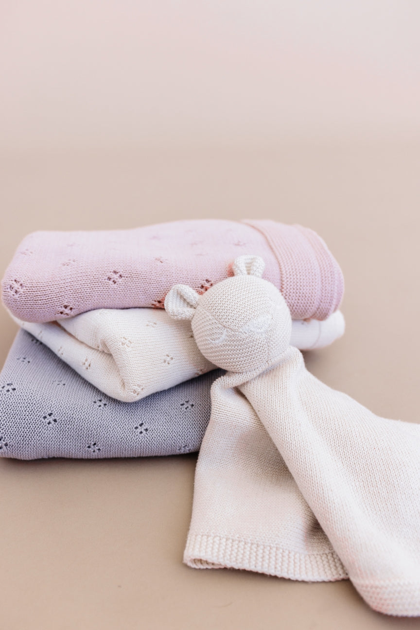 Three folded knitted baby security blankets in pastel pink, white, and light lavender-gray, with a cream knitted bear-shaped teether attached, on a soft beige background.