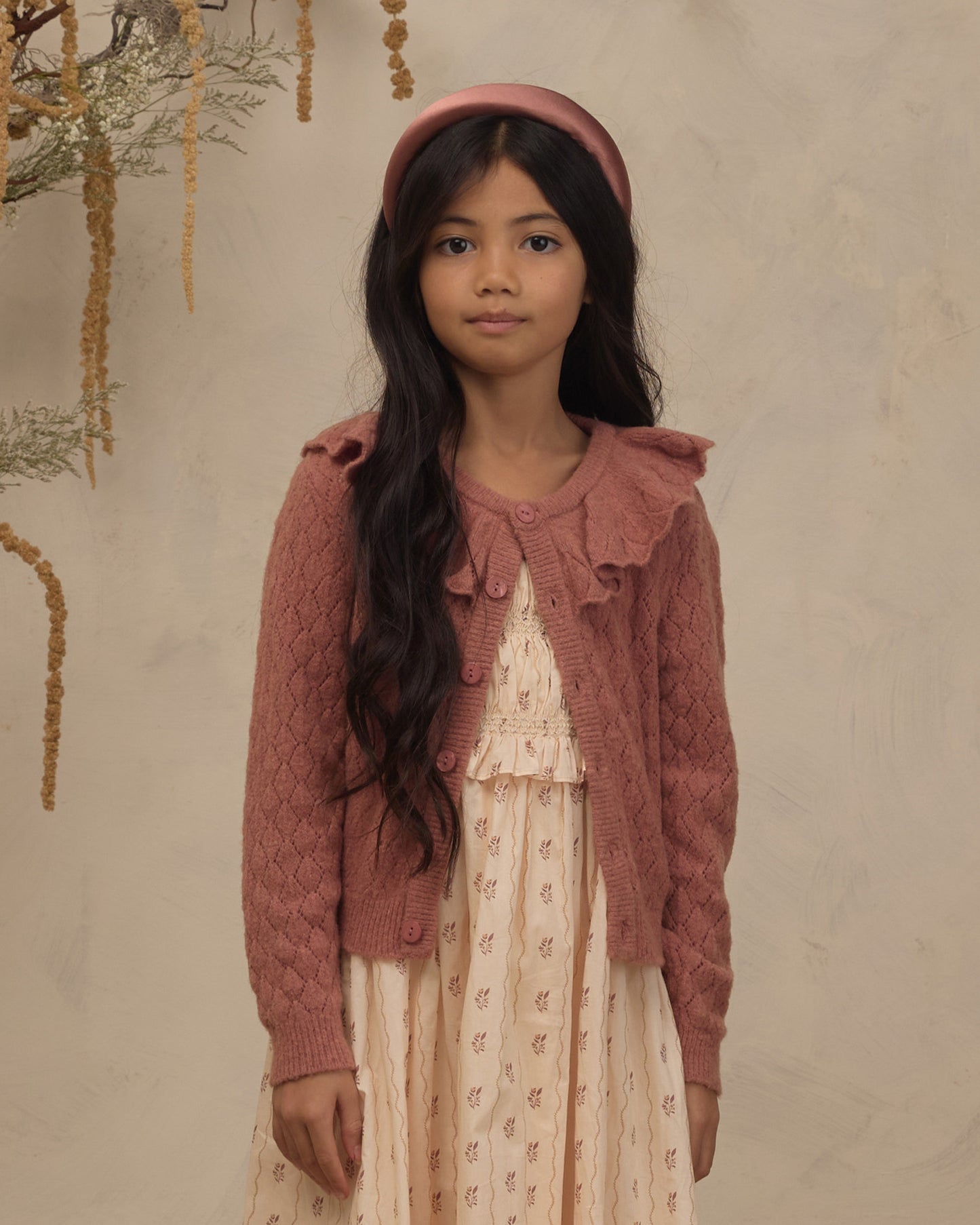 A young girl with long dark hair stands against a textured beige wall wearing a dusty rose knit cardigan over a cream patterned dress, with a matching dusty rose headband.