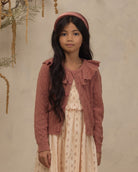 A young girl with long dark hair stands against a textured beige wall wearing a dusty rose knit cardigan over a cream patterned dress, with a matching dusty rose headband.