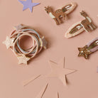 A styled flat-lay of hair accessories on a dusty pink background, featuring a bundle of pastel elastic hair ties with glittery star accents, two deer-shaped hair clips, and two star-shaped hair clips.