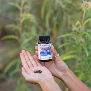Hand holding a bottle of Motherlove More Milk Special Blend with two capsules in front of green foliage.