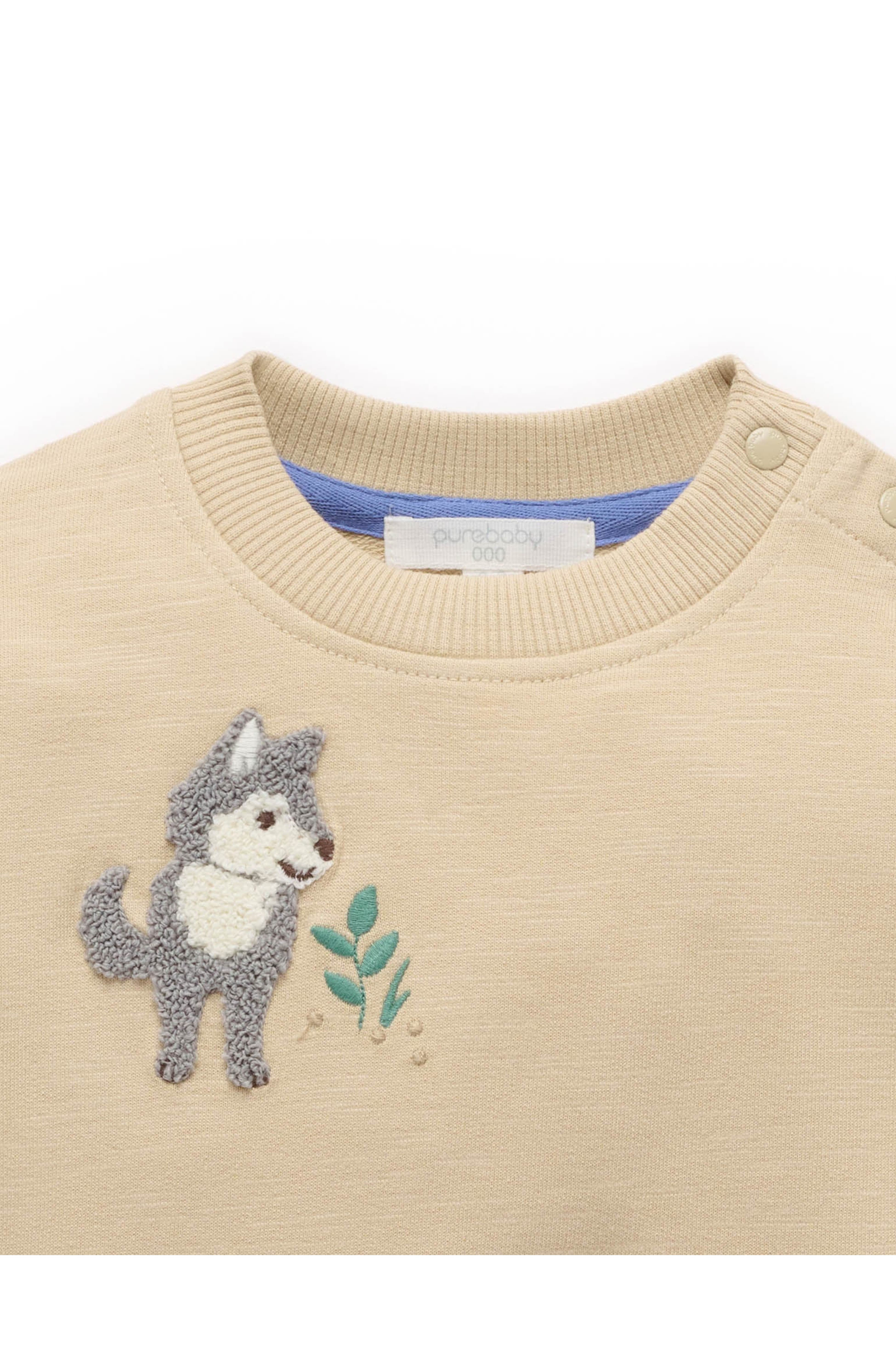 A close-up of a beige knit baby sweater featuring an embroidered gray and white wolf holding green leaves on the chest.