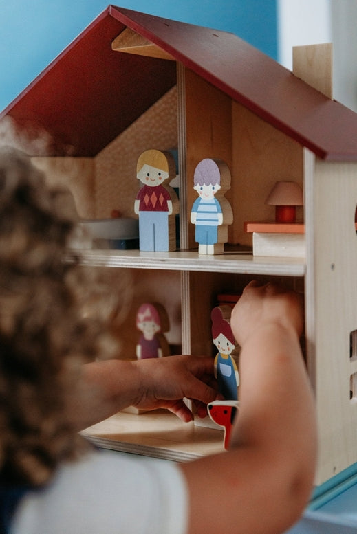 A young child playing with an open-faced wooden dollhouse by Mentari, arranged on a white table with miniature wooden furniture and small figurines.