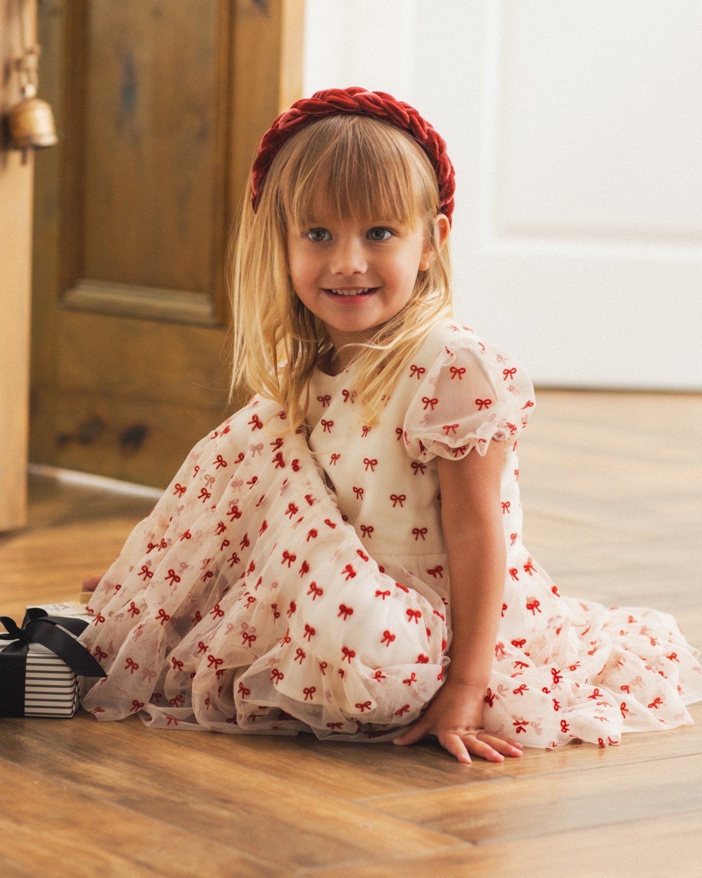 A young girl with blonde hair sits on a wooden floor wearing a white tulle dress with a red bow pattern and a red braided headband. A small striped gift box is next to her.