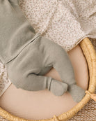 A close-up of a baby's foot wearing a sage green waffle-knit bootie, resting on a beige mattress inside a woven rattan bassinet with a floral blanket.