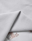 A close-up of a soft, light gray fleece blanket with a folded corner revealing a white underlay printed with a small pink spotted animal motif.