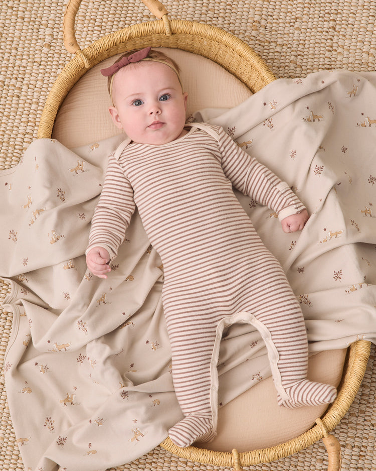 A baby lying on their back in a woven bassinet, wearing a beige and white striped footed sleeper and a dusty rose headband, surrounded by a matching beige blanket with a small floral print.