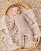 A baby lying on their back in a woven bassinet, wearing a beige and white striped footed sleeper and a dusty rose headband, surrounded by a matching beige blanket with a small floral print.