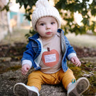 Baby sitting outdoors wearing a denim jacket, yellow pants, and a white knit hat.