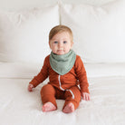 Baby wearing a rust-colored outfit with a green bib on a white blanket