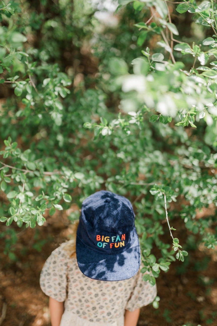 A child viewed from behind wearing a navy blue tie-dye baseball cap with colorful embroidered text that reads 'BIG FAN OF FUN', standing outdoors in front of leafy green trees.