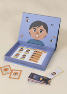 An illustrated open storage box or game box on a light beige background, containing printed cards and small cards placed around it. The box lid shows a cartoon face with eyes, mouth, and hair, and the inside contains labeled cards with matching facial features.