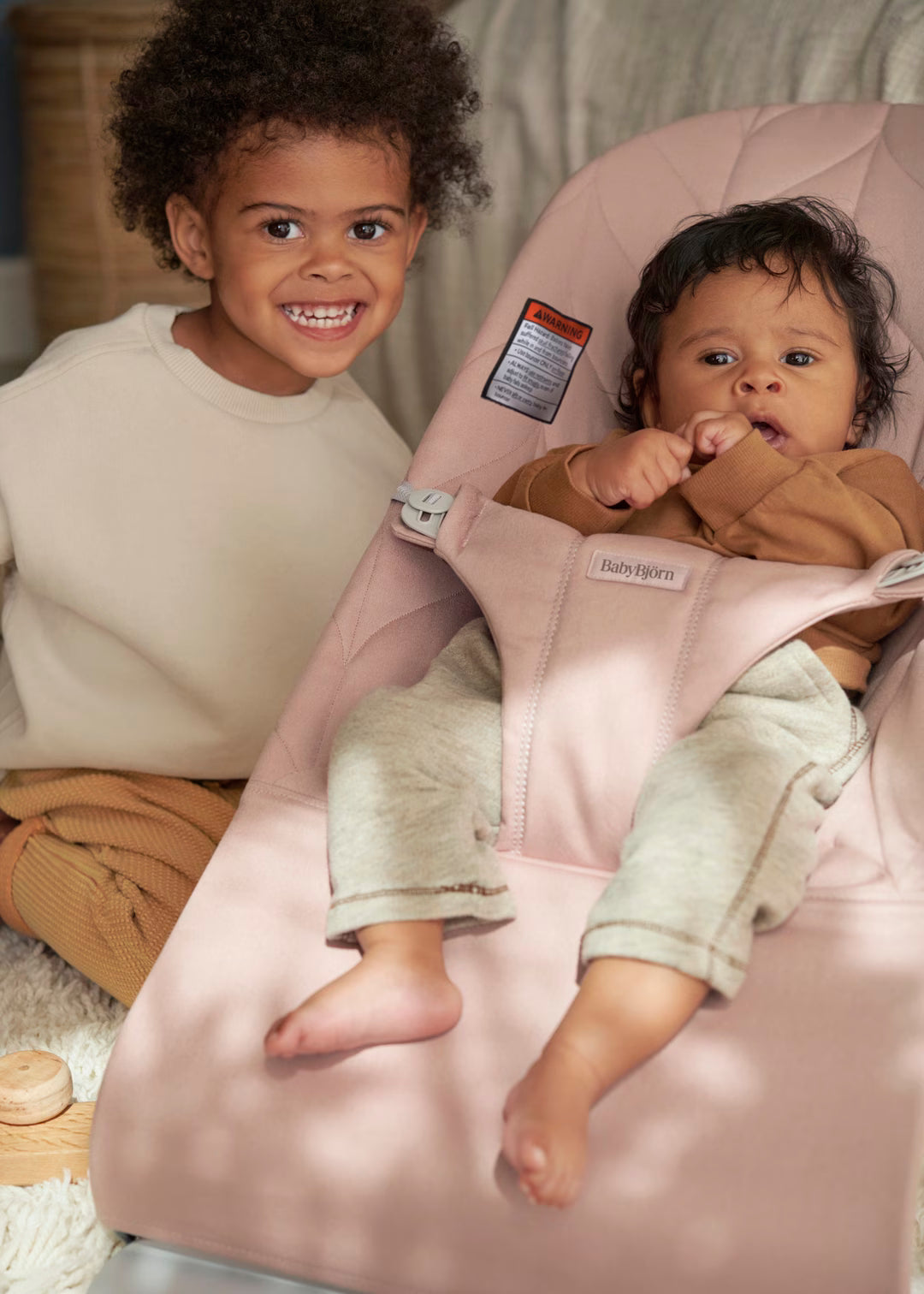 Child sitting on a pink baby bouncer with another child standing nearby.