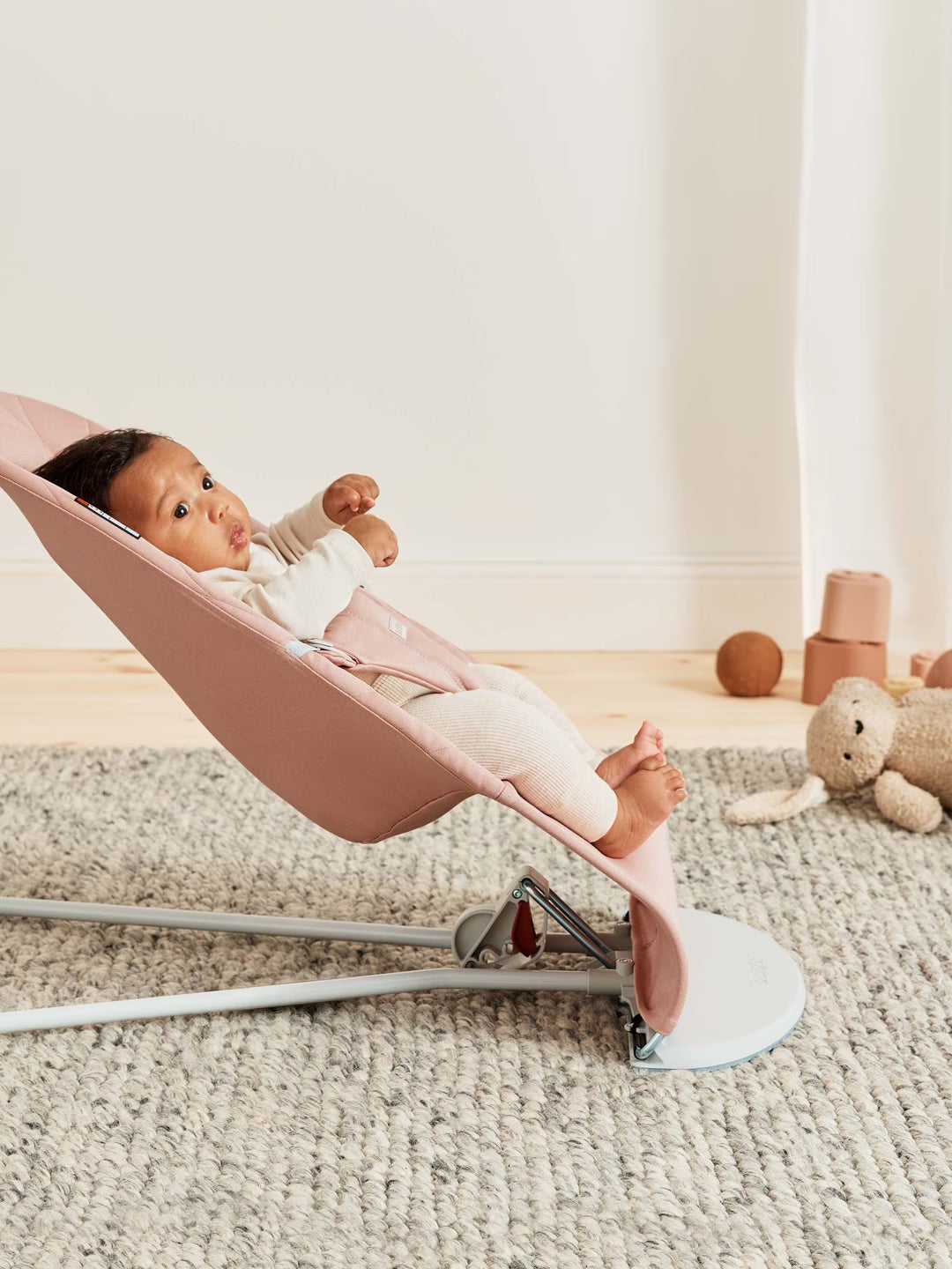 Baby in a pink baby bouncer with toys on a carpeted floor
