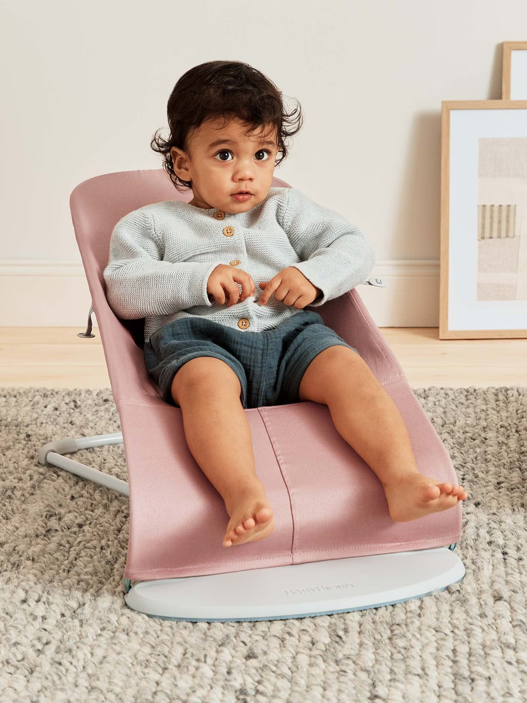 Child sitting on a pink baby bouncer in a room with a light-colored wall and framed picture.