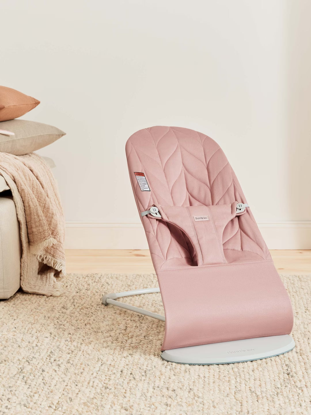 Pink baby bouncer on a beige carpet with a neutral-colored wall and pillows in the background.