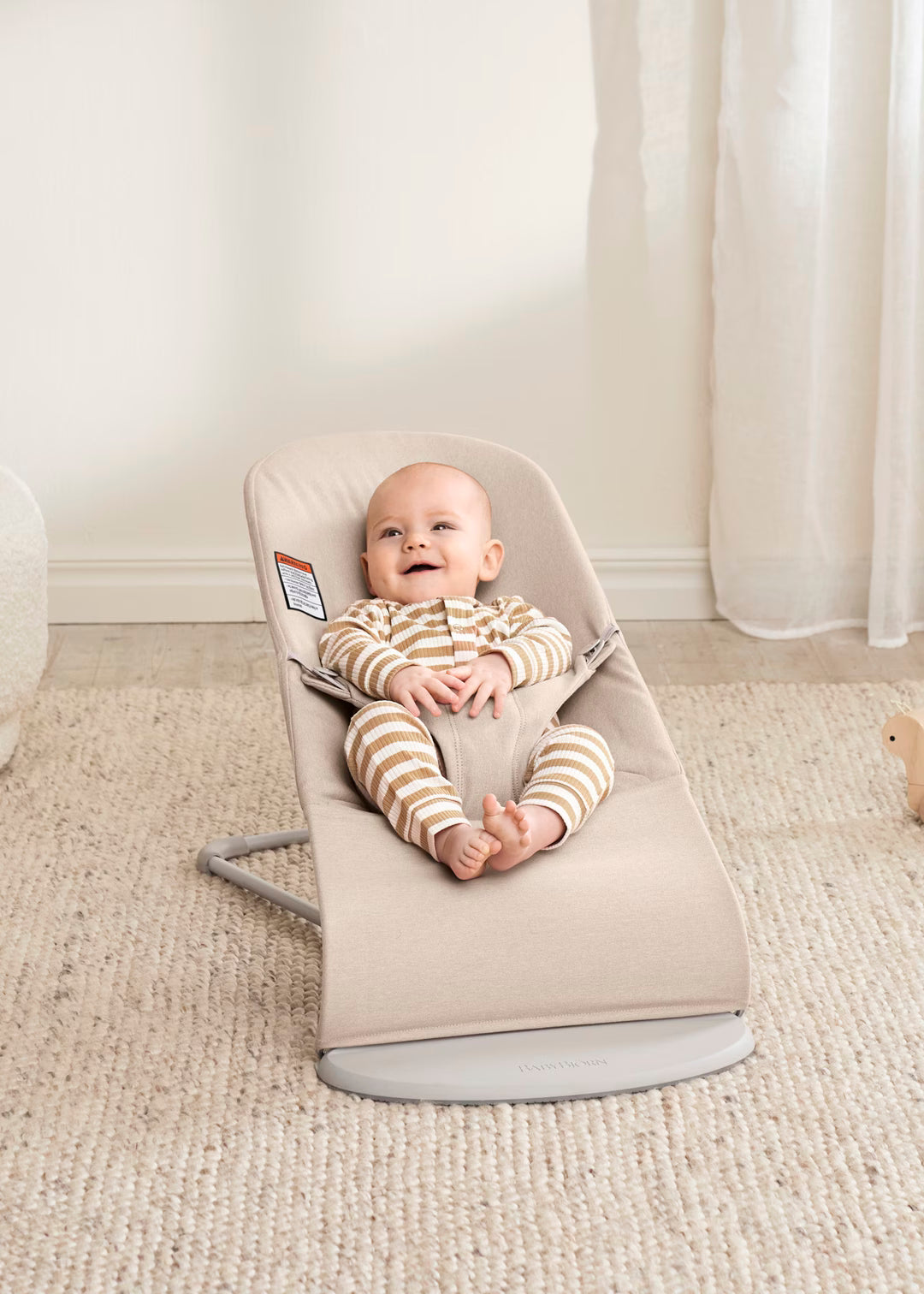 Baby in a beige baby bouncer on a carpeted floor with white curtains in the background