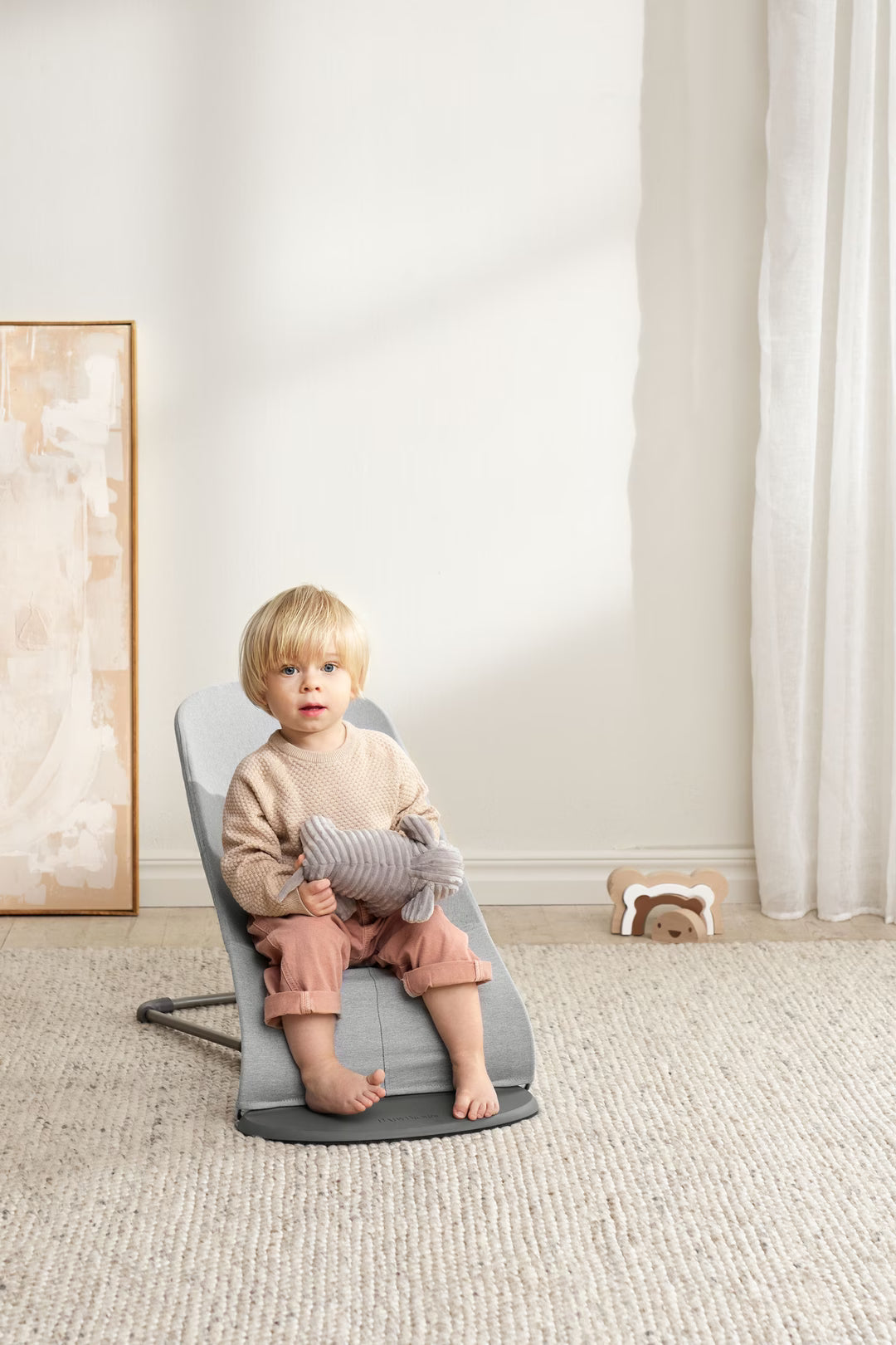 Child sitting on a gray chair in a room with white walls and a window.