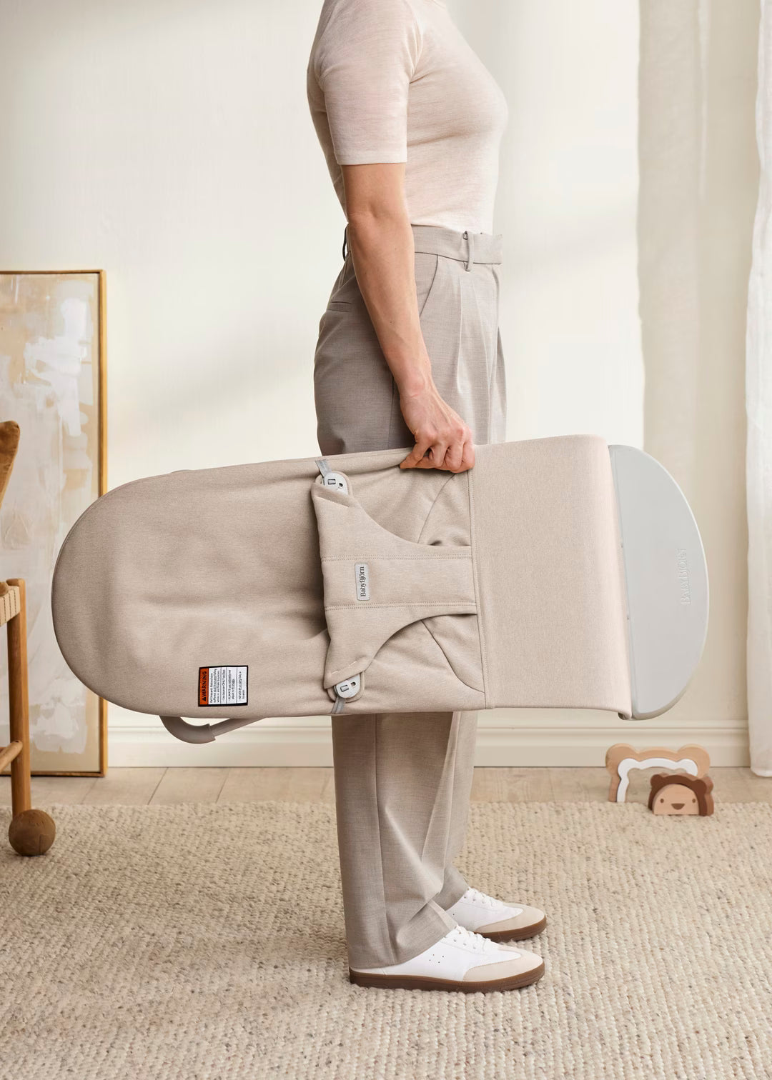 Person holding a beige baby bassinet in a room with neutral decor
