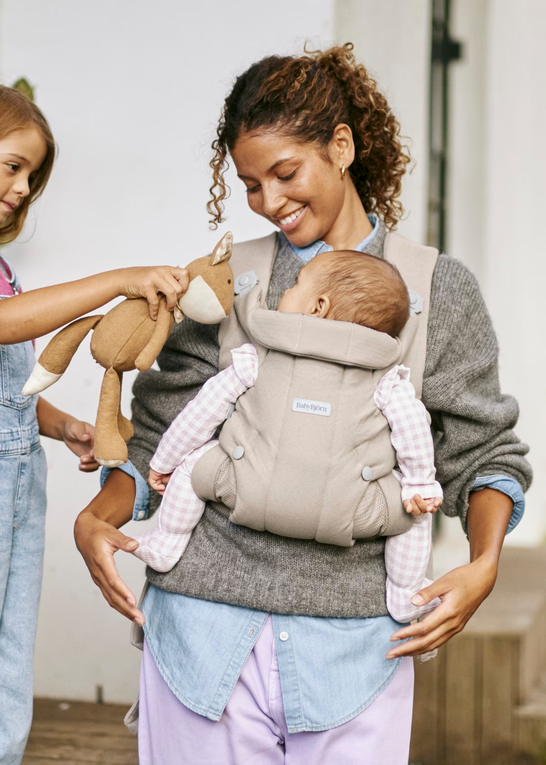 A woman wearing a gray knit sweater and light blue shirt holds a baby in a beige KAVU baby carrier. A young girl on the left holds a brown plush toy.