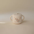 A small cream-colored ceramic mug with two side handles and a lid, decorated with a delicate pink and green floral pattern, set against a solid beige background.