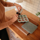 A person pouring liquid from a metal pitcher into a green silicone baby food freezer tray on a wooden countertop, with a matching tray and lid set nearby.