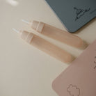 Two beige lip gloss tubes on a light pink surface with a small notebook in the background.