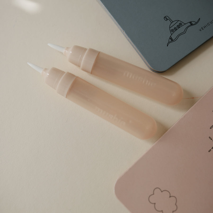 Two beige lip gloss tubes on a light pink surface with a small notebook in the background.